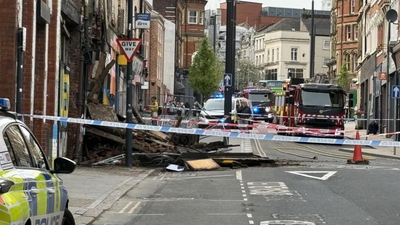 A partially collapsed building in Leeds is taped off by police and fire crews, with emergency workers at the scene. Two police vehicles and two fire engines are in shot. Building debris spills across a pavement and a road. 