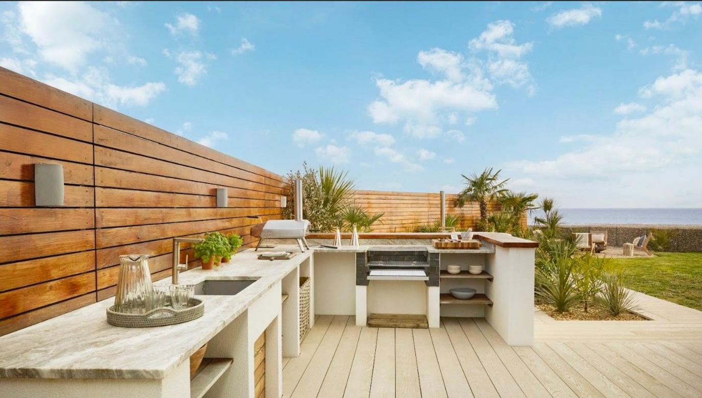 An outdoor kitchen with marble work tops, a pizza oven and sink. It sits on top of a large decked area and is surrounded by wooden panelling and lights.