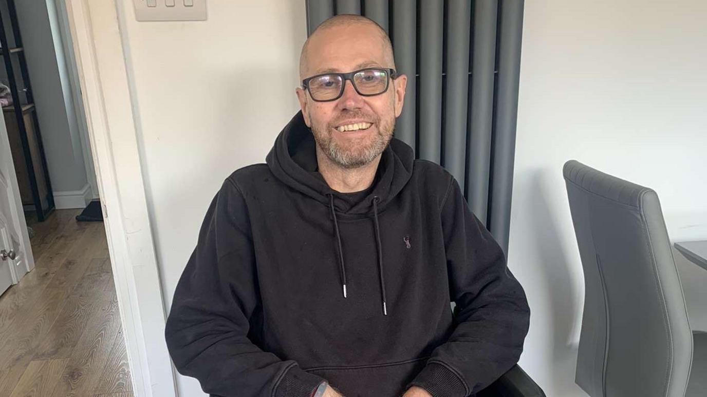 Nick Ball wearing a black hoodie and black framed glasses. He has a bald head and a closely shaven beard. He is sitting down in a white living room beside a grey dining chair. He is smiling at the camera, but looks weak from cancer treatment.