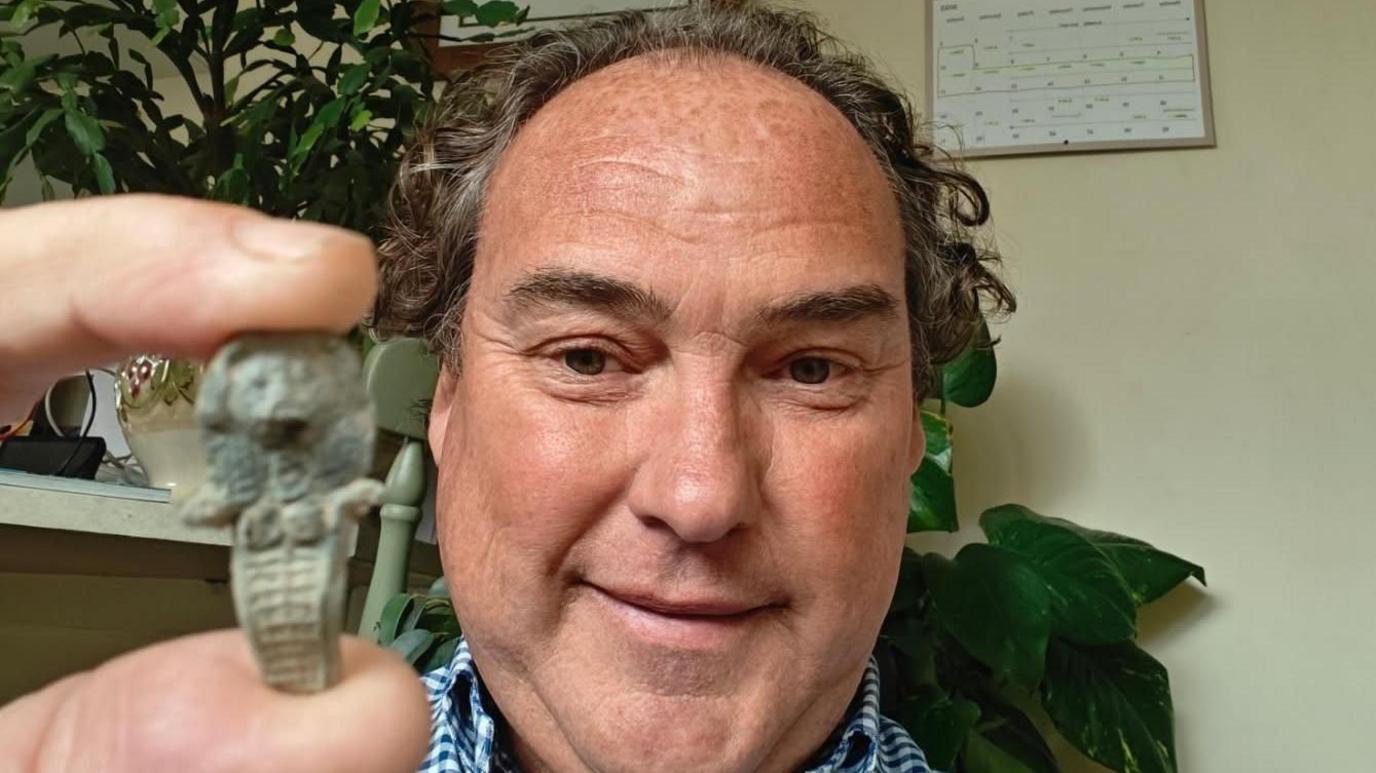 Mark McMullan has taken a selfie while holding a medieval doll between his thumb and forefinger. He is smiling into the camera next to the statue. Is has ribbed legs and the look of an Egyptian mummy.