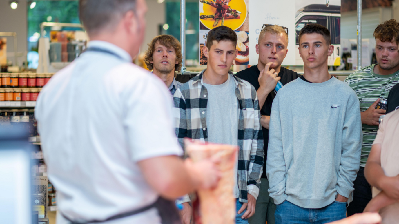 Five men watching a man show them how to butcher a piece of meat in a food shop