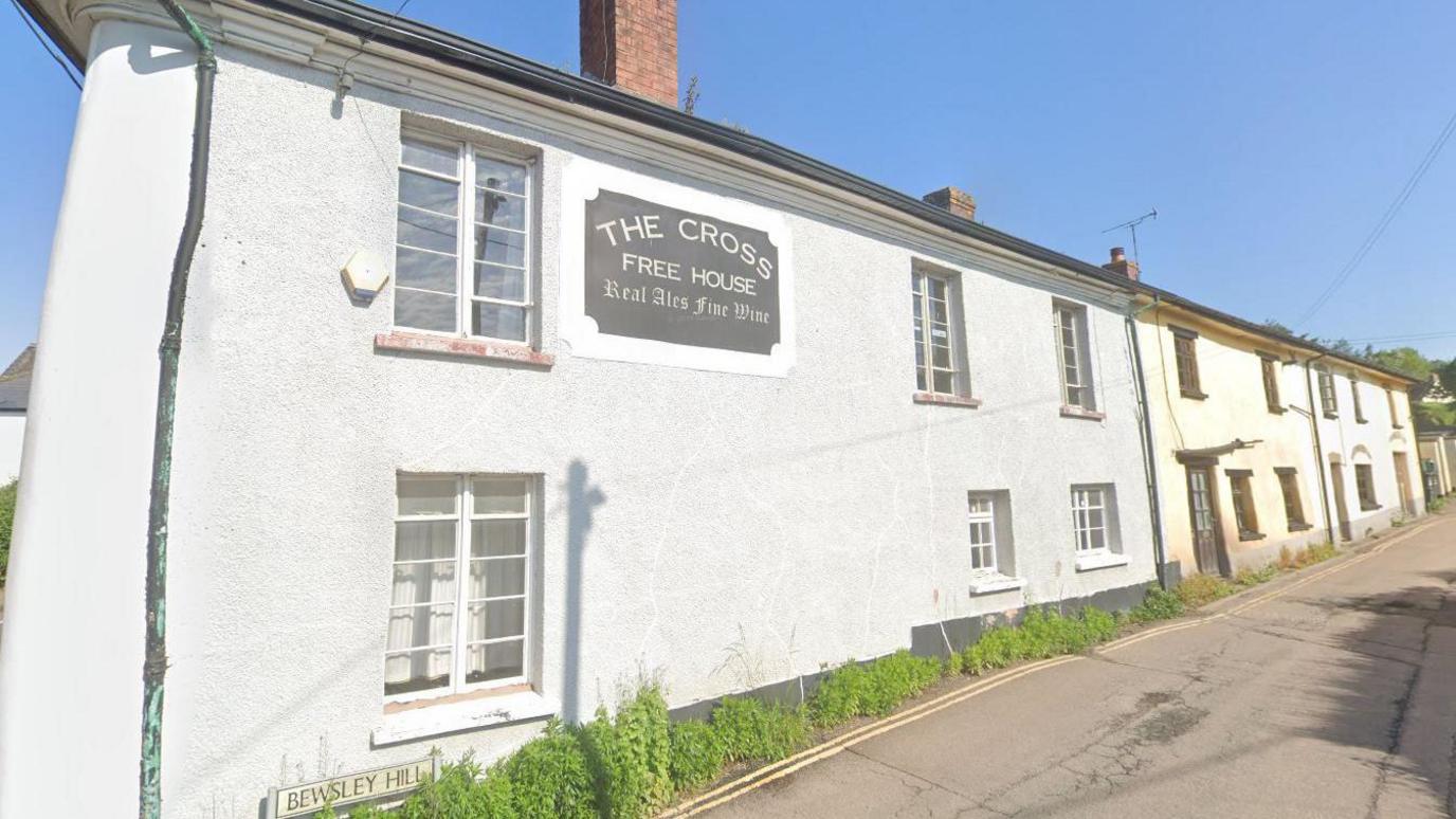 Google Street View image of The Cross Inn in Copplestone on a sunny day. The building has grey walls and has two other buildings attached to it. A sign saying "The Cross Free House - Real ALes and Fine Wines" is on the wall.