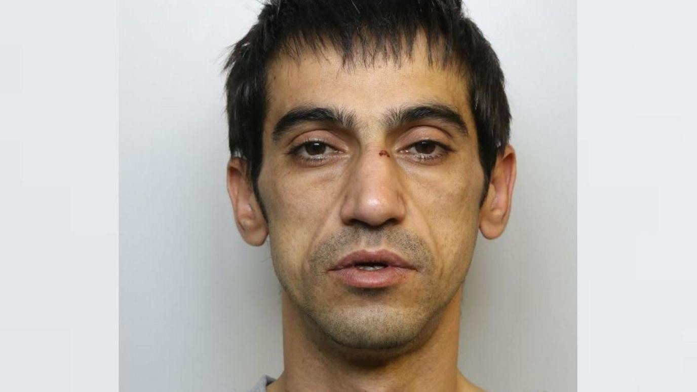 A mugshot of a thin man wearing a grey jumper. He has dark hair, stubble and a cut on his nose.