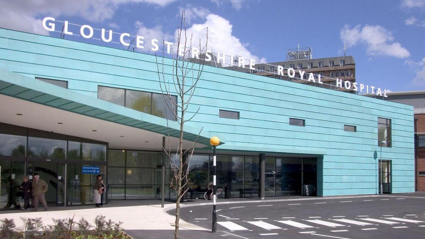 The front exterior of Gloucestershire Royal Hospital. It is a large rectangular building clad in blue panels. On the left there is a sheltered area, and a zebra crossing heading. It is a sunny day with wispy clouds in the sky. 