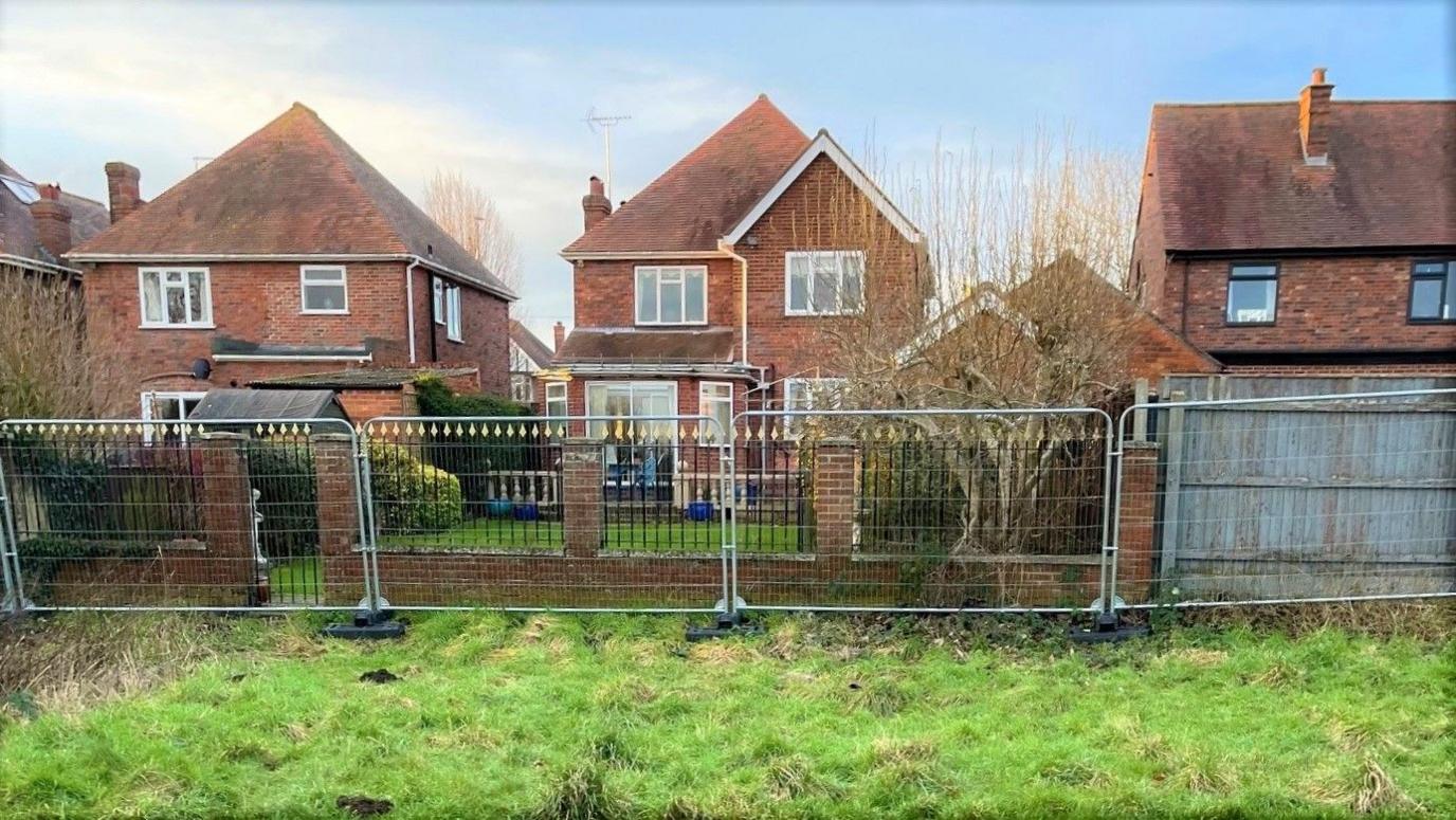 A view from inside the field towards a row of three red brick detached houses.  Heras fencing has been installed along the perimeter line of their back gardens, running parallel to a low garden wall. 