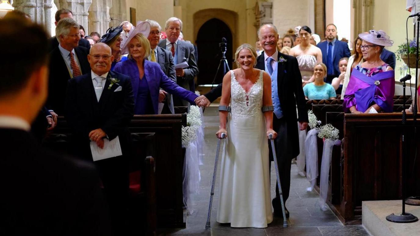 A blonde woman in a white wedding dress makes it down the aisle on clutches. 