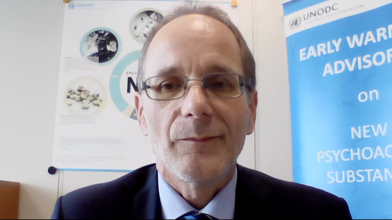 Martin Raithelhuber faces the camera while sat in an office, wearing a navy blue blazer, pastel blue shirt and navy-blue, striped tie and glasses. Behind him are two unfurled banners: one to his left is referring to an early warning advisory message from the United Nations Office on Drugs and Crime (UNODC) about new psychoactive substances (NPS); over his right shoulder the second banner describes the effects of NPS.