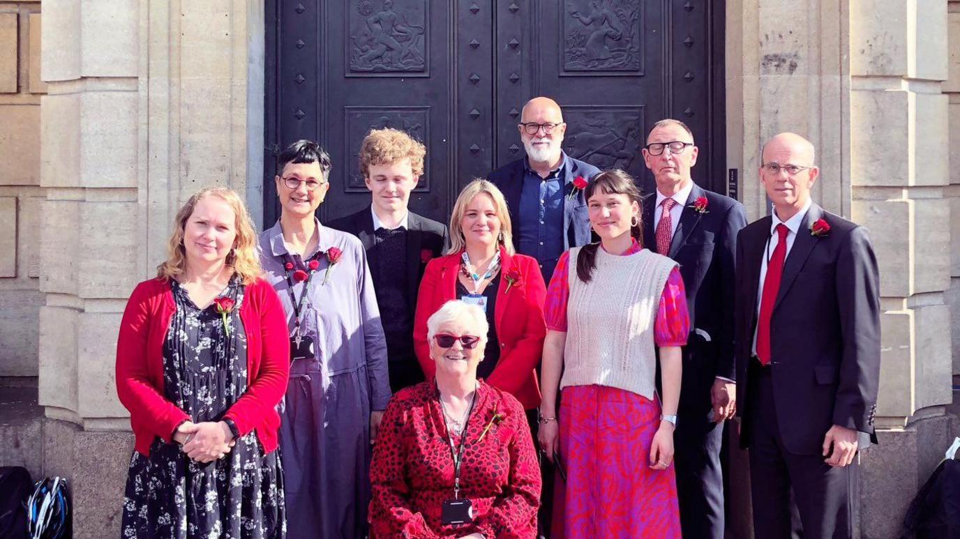 9 people pose in front of a grand door at the exterior entrance to the Cambridge Guildhall.