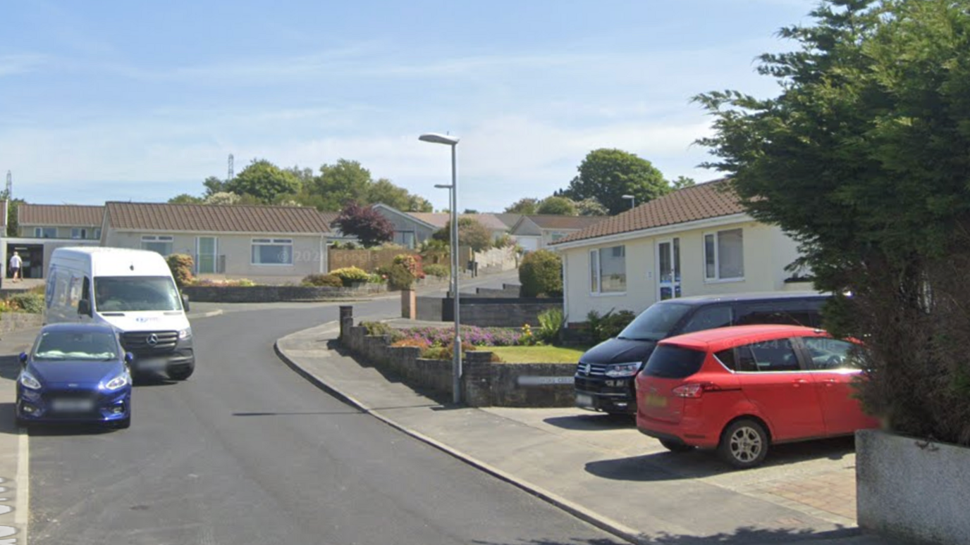 A street view of a residential road in Cornwall. There are houses on either side of a road which has cars parked along it.
