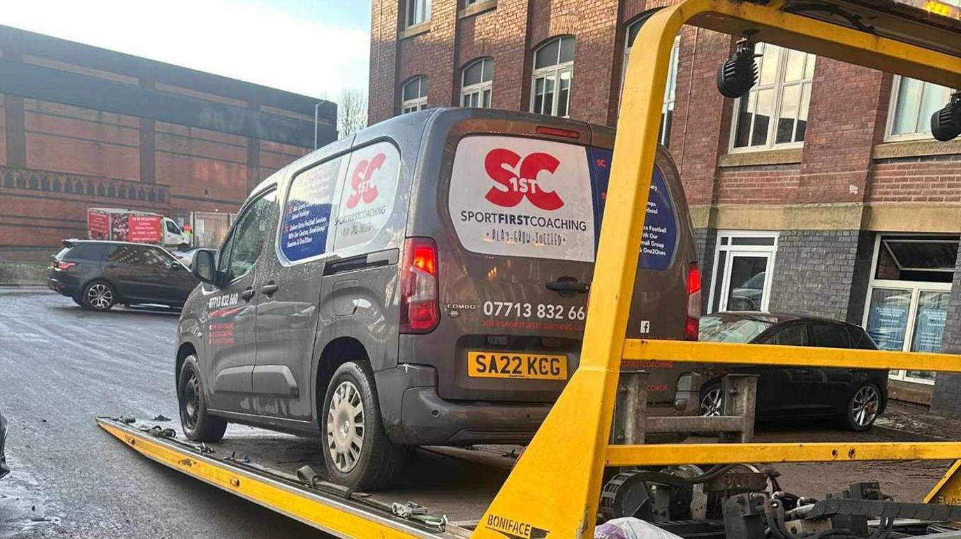 A grey van advertising sports coaching services being hauled onto a ramp and taken away outside Meadow Mill apartments in Stockport. 