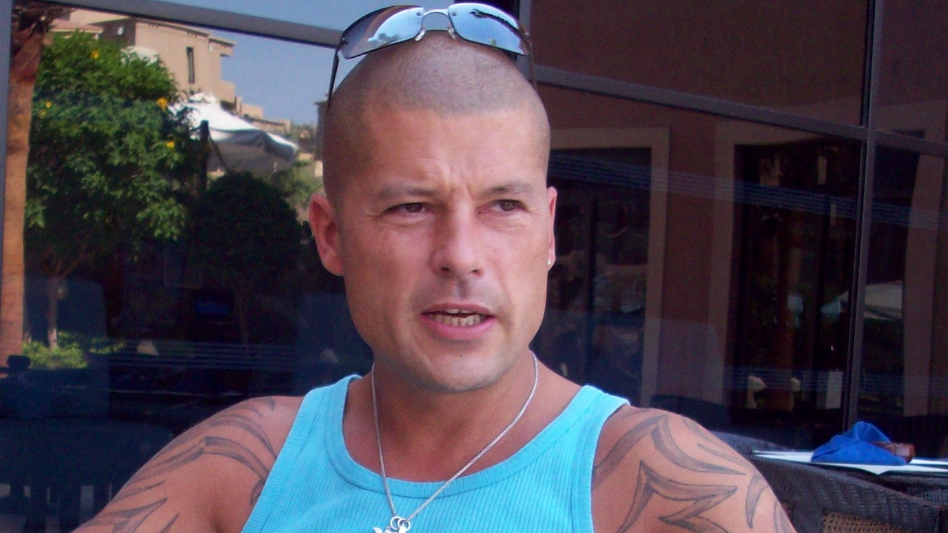 Mr Arter is looking to the right of camera, wearing a blue vest top, silver necklace and has sunglasses resting on top of his shaven head.