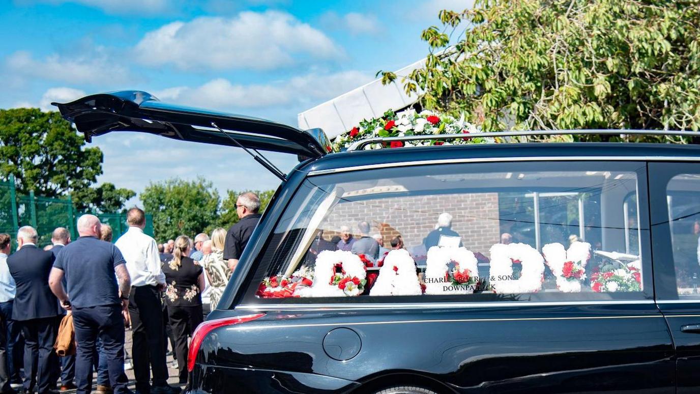 A hearse is viewed from the side with white flowers spelling out daddy in capital letters. A number of mourners are standing to the side of the hearse