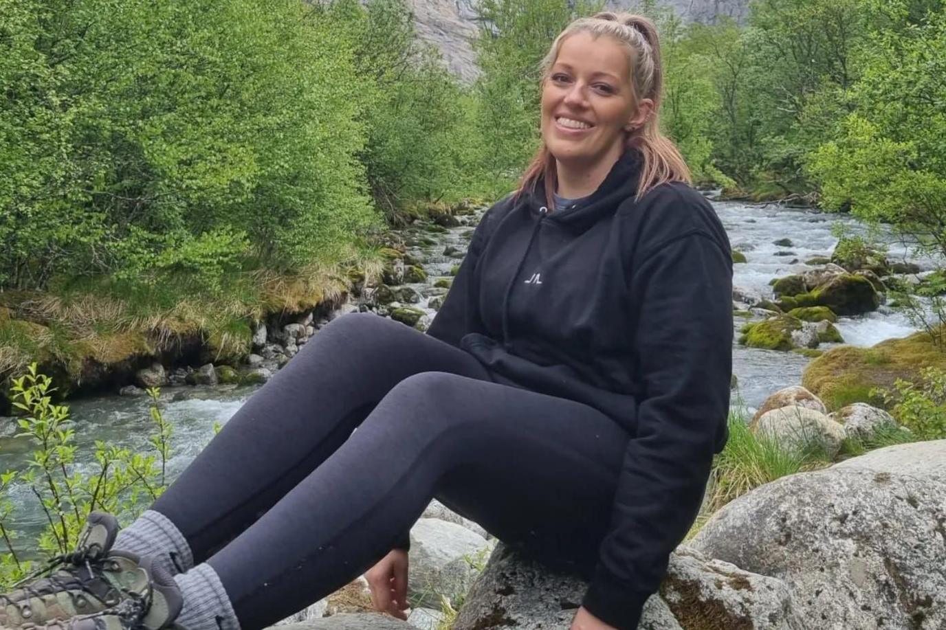 A young woman sits on a rock next to a river with lush greenery in the background. She is smiling into the camera with her hair pulled back, wearing a black hoodie, black leggings and walking boots