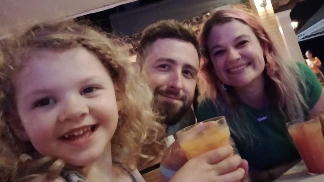 A young girl with curly blond hair smiles at the camera. Her parents are behind her, a man with a brown beard and short hair, and a woman with long, light-pink hair. They are both smiling, and appear to be in a restaurant.