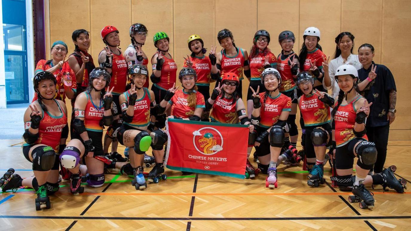 A Chinese Nations Roller Derby team photo, with some of the members making the peace sign. Two of the members on the first row are holding the team flag, which depicts a golden dragon on a red canvas. Everyone is wearing their kit and is smiling for the camera.