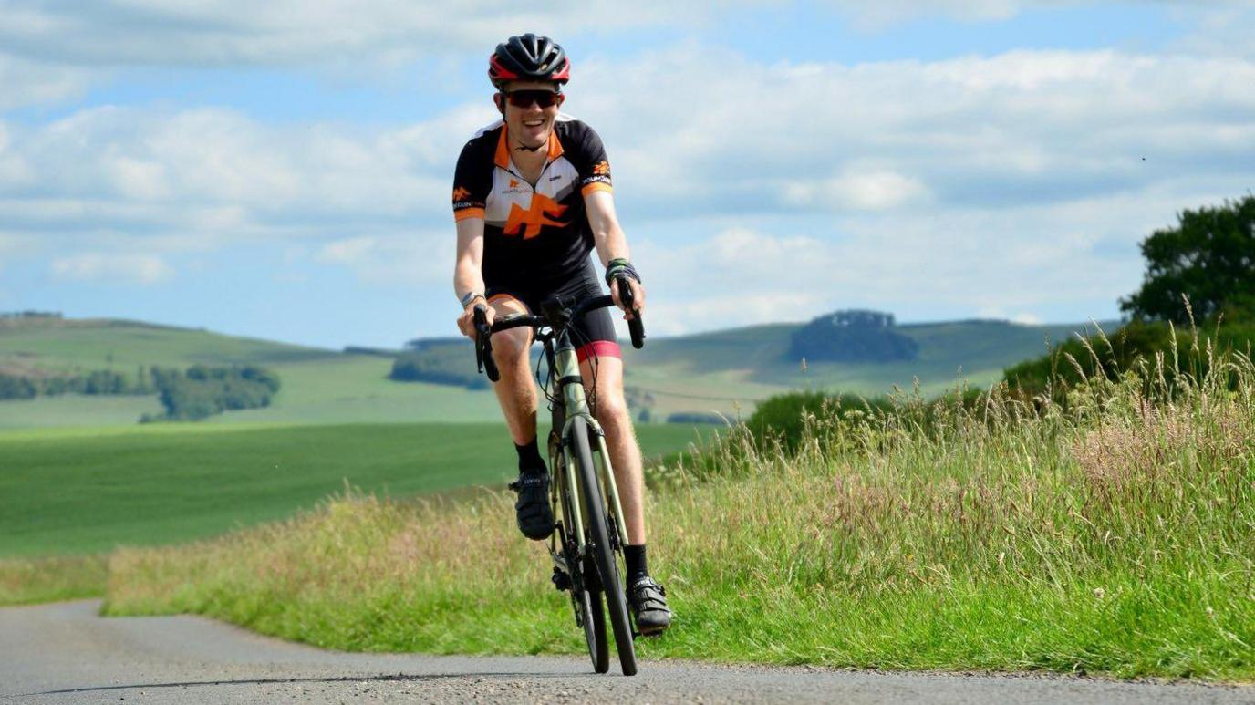 Alex Staniforth riding a bike. He is wearing a black helmet and outfit as well as sunglasses. He is smiling as he rides towards the camera. Green fields can be seen in the background.