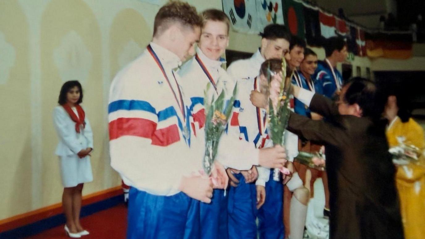 Four Team GB swimmers stand in the podium in a line as they are given bronze medals by a dignitary at the 1998 Seoul Paralympics, while a woman looks on. The four are all wearing blue, red and white Team GB track suits and holding flowers. Mark, who has close-cropped blond hair, is second from left and is smiling into the camera.