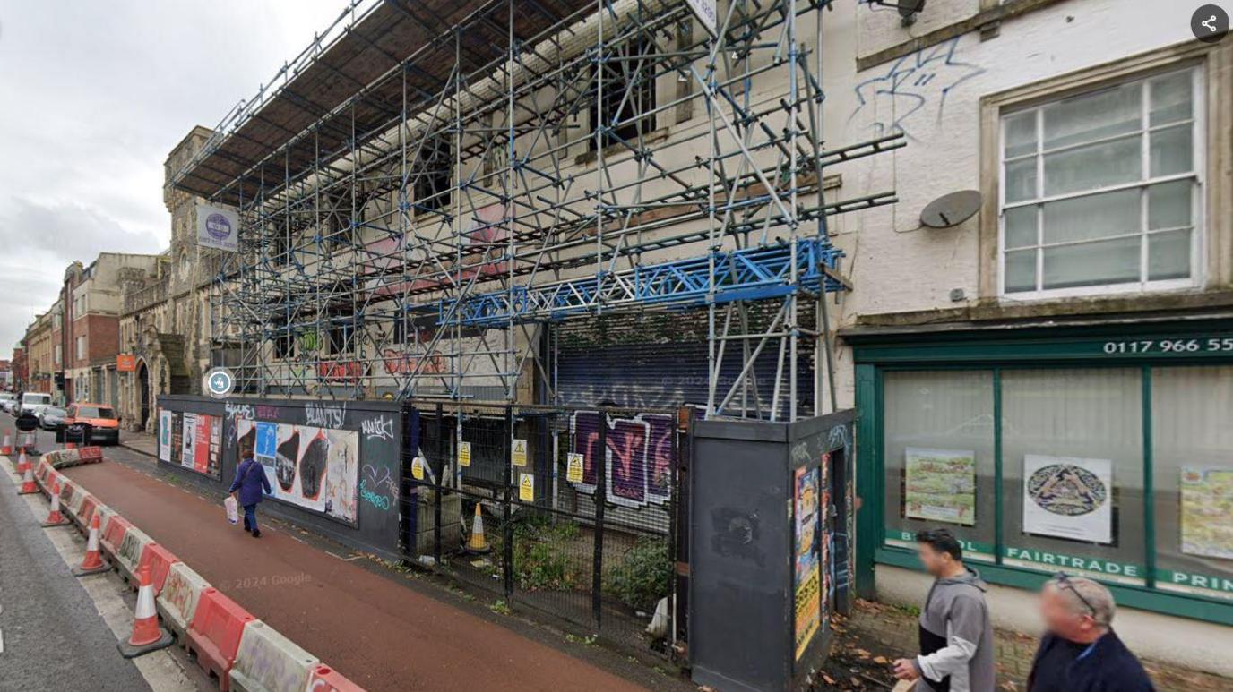 A building on Bedminster Parade in Bristol which is covered in scaffolding. There is graffiti on it and a fence blocking access to it. Hoarding covers the pavement and pedestrians are walking in the bus lane, which is coned off.