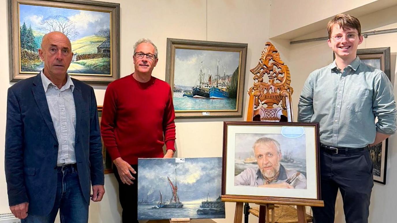 Michael Rigg, wearing a blue suit and white shirt, and Ian Rigg, wearing a red jumper and dark trousers, stand among an exhibition of paintings by  their father, Jack Rigg, whose portrait is also shown. To the right stands art expert Dominic Cox, who wears a blue shirt and dark trousers.