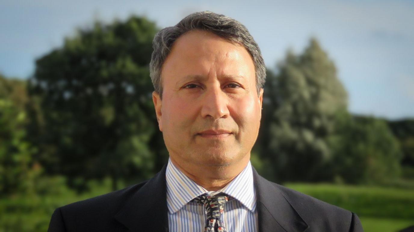 Rakesh standing outside, trees in the background, the sun is shining. He is wearing a dark grey suit, yellow and blue striped shirt and a tie with elephant pattern