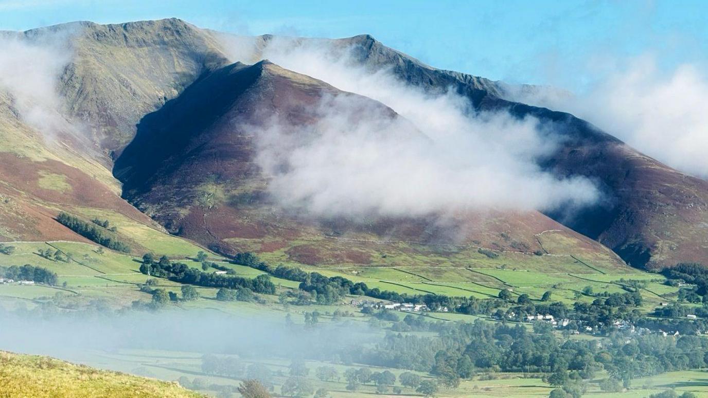 Belcathra which has steep sides and is surrounded by mist. The sky is blue and clear aside from the mist and houses can be seen in the valley below along with trees and fields.