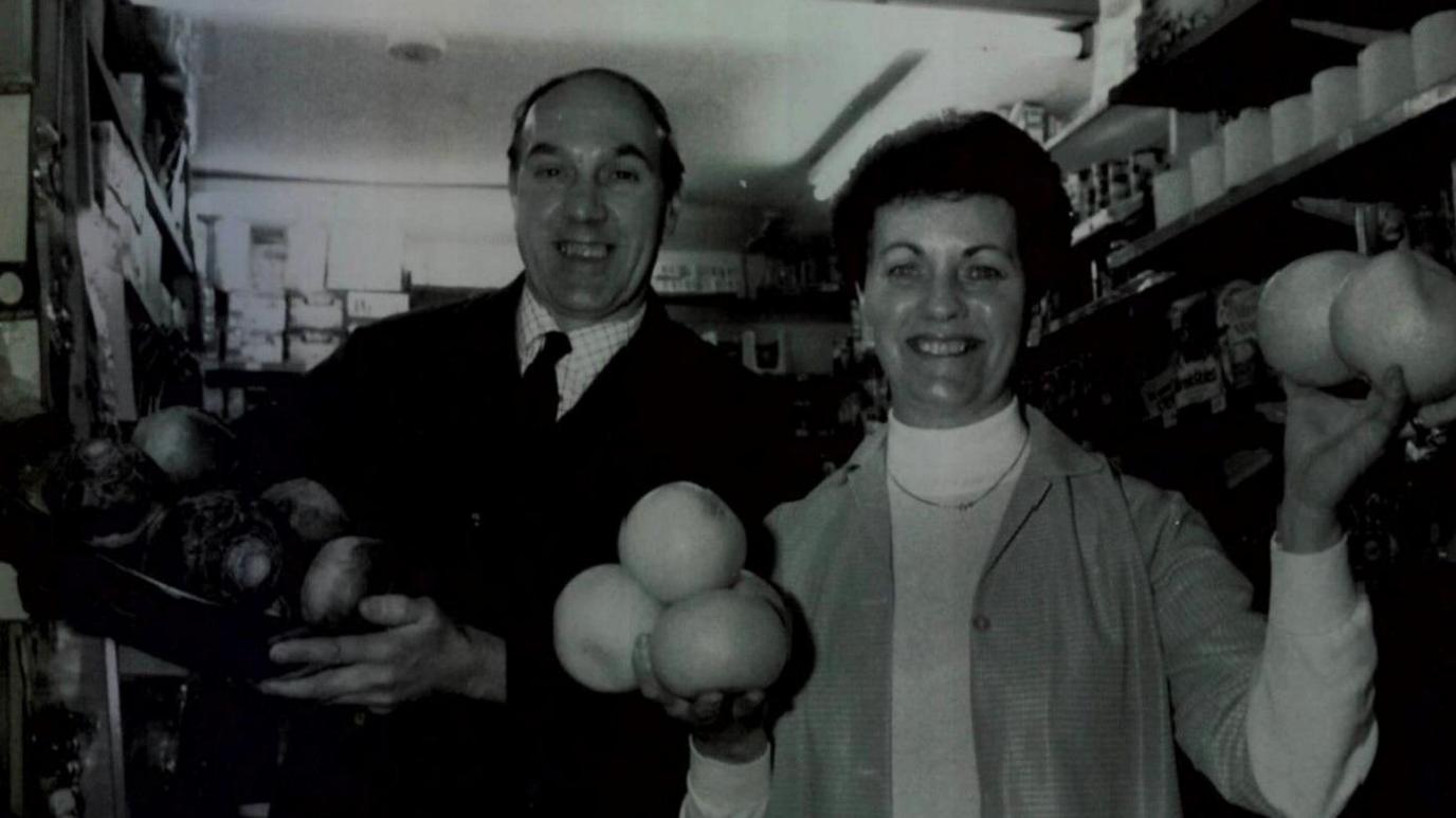 A black and white photo of a man, on the left, and a woman, on the right, holding up fruit and veg and grinning. They are in a shop, surrounded by shelving. He is wearing an overcoat, checked shirt and tie, and she has an over-shirt over a polo-neck. 