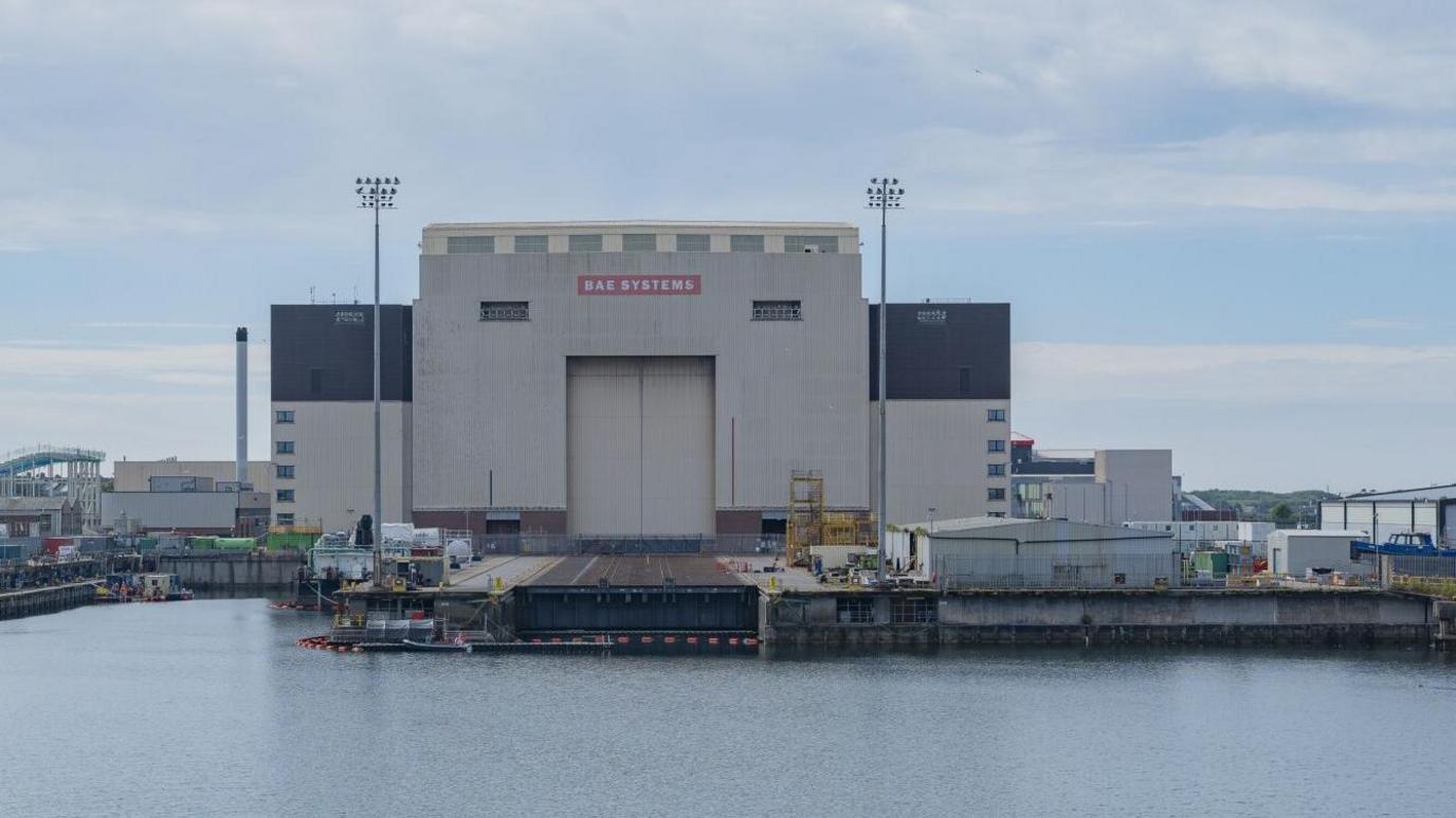 A side view of a large hanger that makes up part of the BAE Systems nuclear submarine shipyard in Barrow-in-Furness. The hanger backs onto a small port of water.