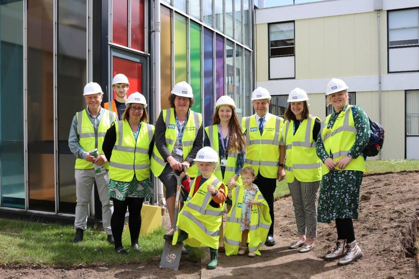 A team of people dressed in hard hats and luminous vests stand on the bare earth and grass in a hospital courtyard. Chief executive Alice Webster is holding a spade.