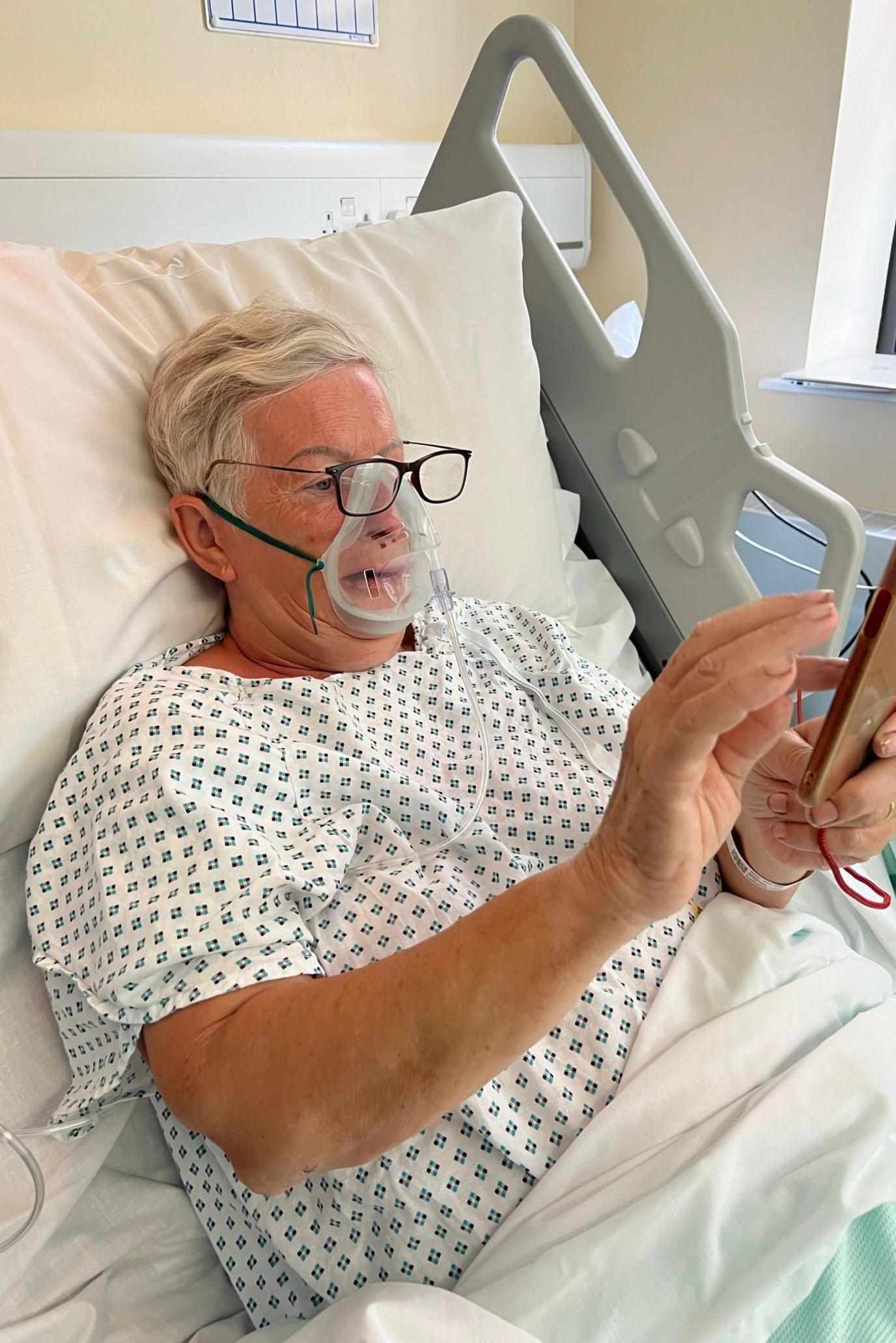 A woman with short white hair sits up in a hospital bed, wearing a hospital gown. She is wearing an oxygen mask and glasses and views content on her mobile phone.