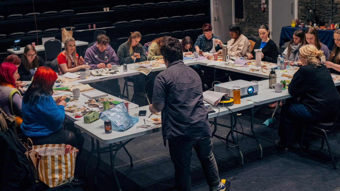 A workshop with a group of young people all sat around long tables grouped in a square. There are water bottles, mugs, books and print outs scattered across the tables with a projector facing away from the shot.