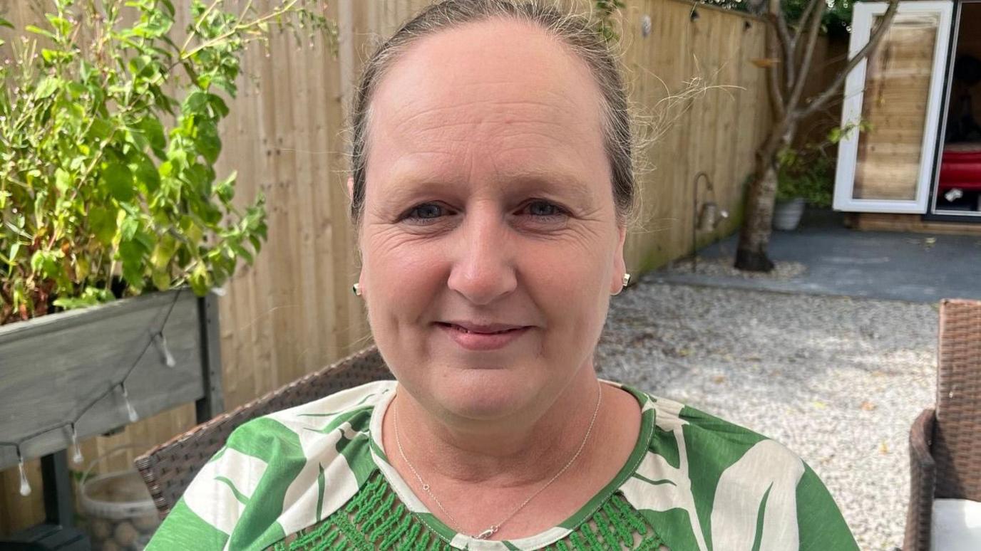 Headshot of Gina Walker smiling into the camera with her hair tied back, a green top with white flowers on it in a garden