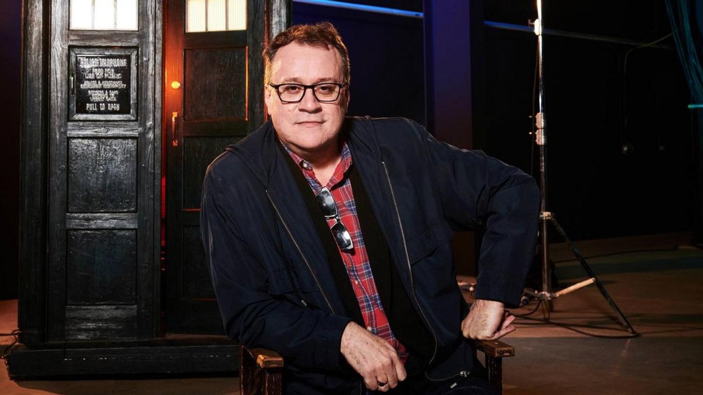 In this photograph Russell T Davies is wearing a red and blue checked shirt with a black jacket. It's a professional photograph taken on the set of Dr Who. You can see the Tardis in the background. He has dark hair and wears black glasses. 