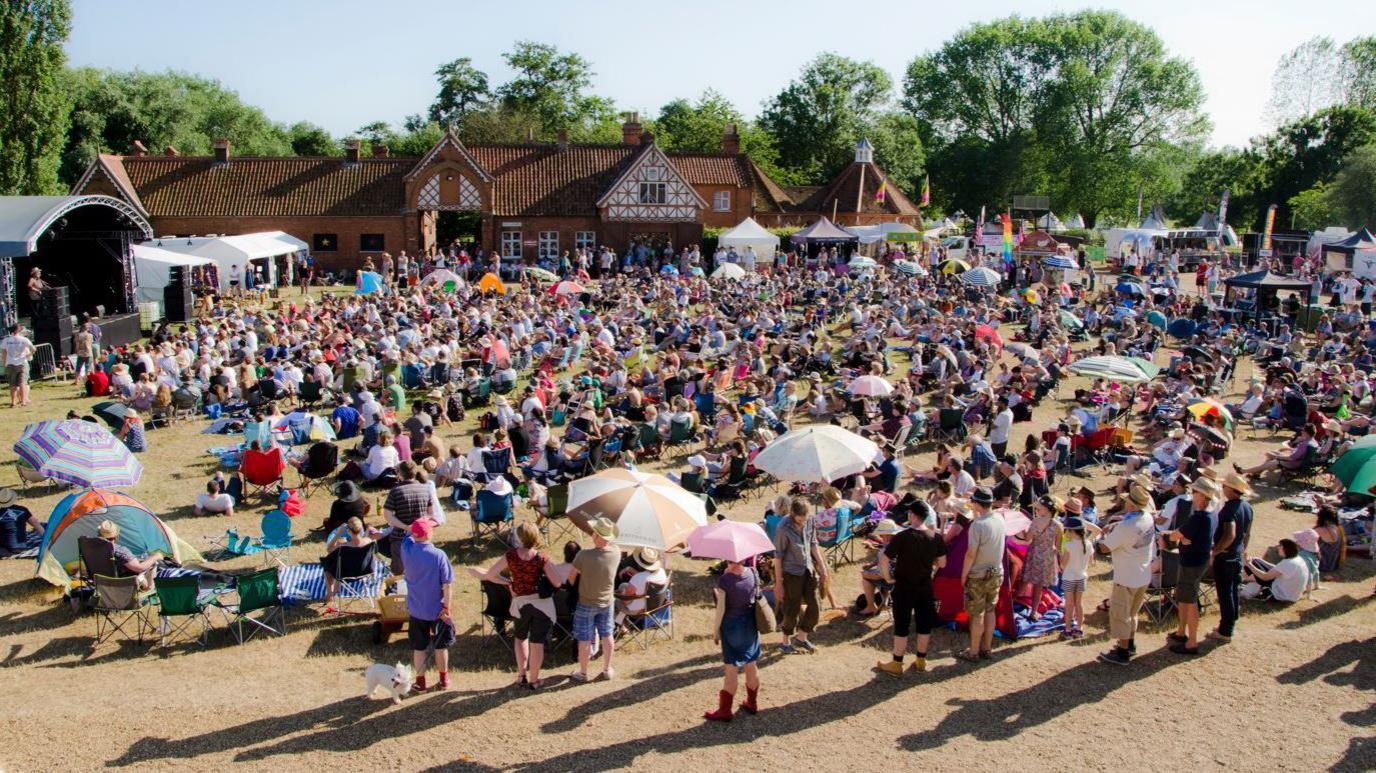 Billy Bragg performing at the Maverick Festival in Suffolk, with a wide shot of the crowd on the grass in front of the outdoor stage and Victorian farm buildings in the background