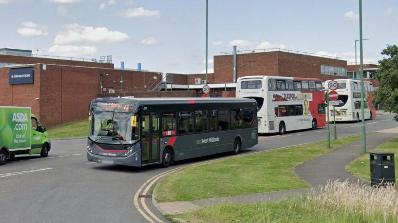 Several buses, some single-deck and some double-deckers, travelling down a road next to some red-brick buildings. There is a bend in the road with a grass verge and footpath to the right. 