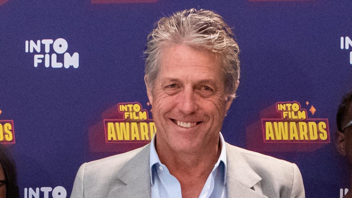 Hugh Grant wearing a light grey suit jacket and a light blue top. He has short light greying hair. He is standing in front of a screen that is purple and says INTO FILM AWARDS in yellow.