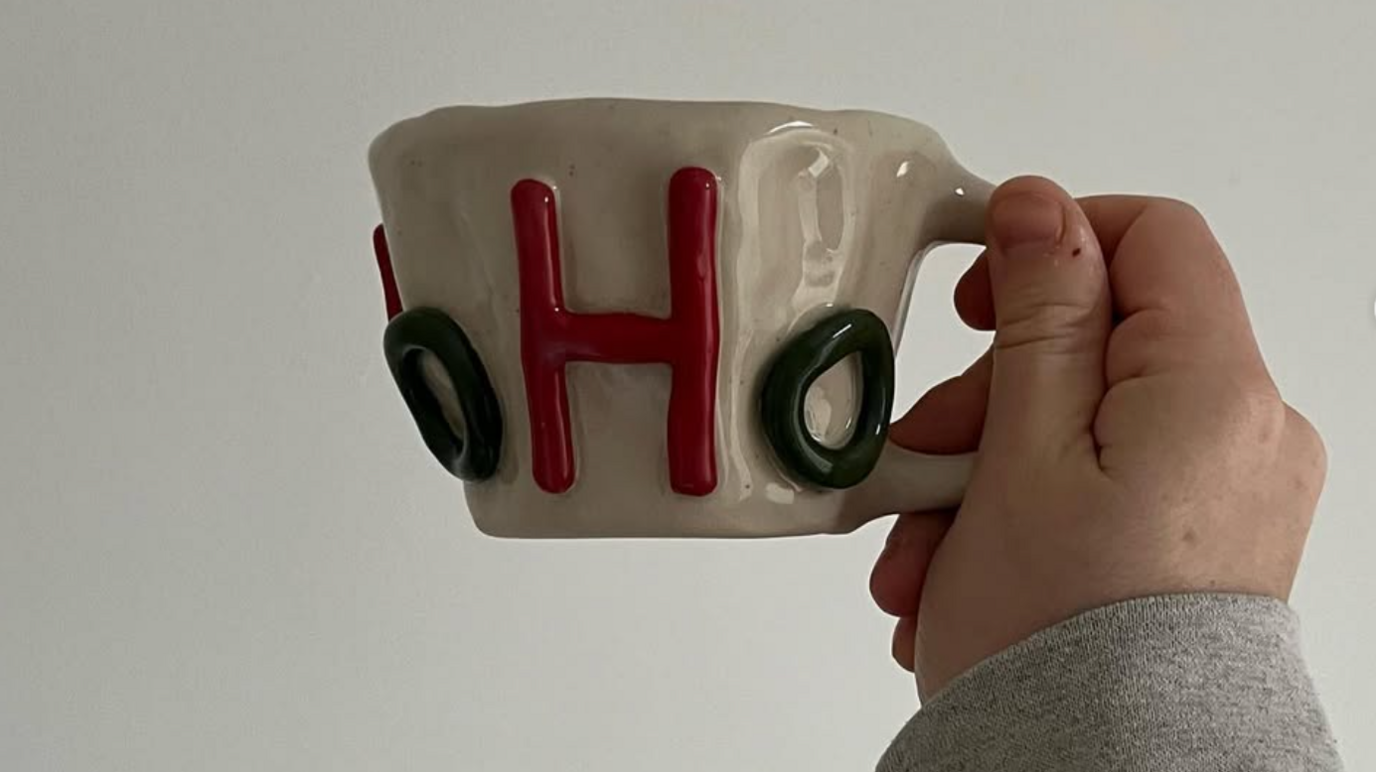 A hand, with a grey sleeve, holds up a mug, beige in colour with a bright red upper case 'H' and a lower case, green 'o'.