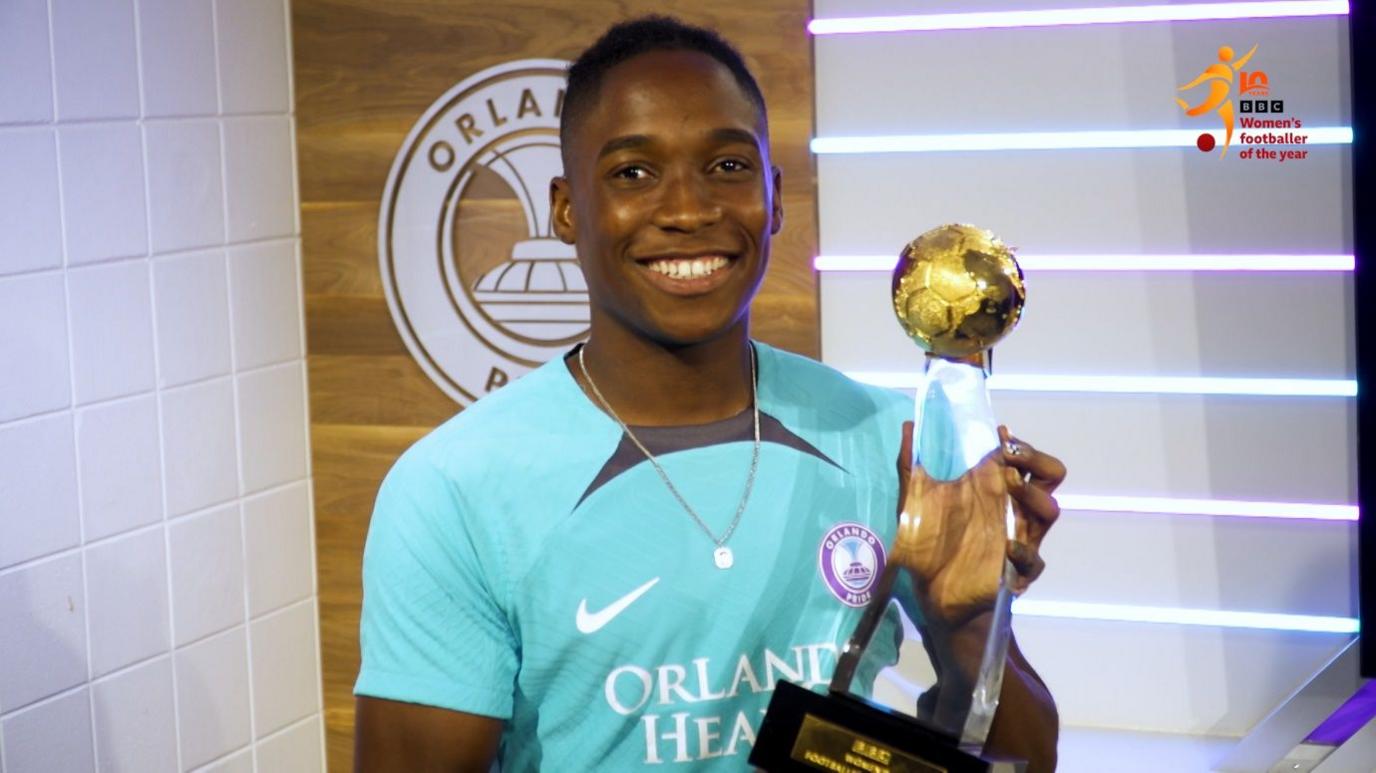 Barbra Banda, who plays for Orlando Pride in the USA, dressed in green training top, smiles as she holds up the trophy for this year's BBC Women's Footballer of the Year in front of her club's logo