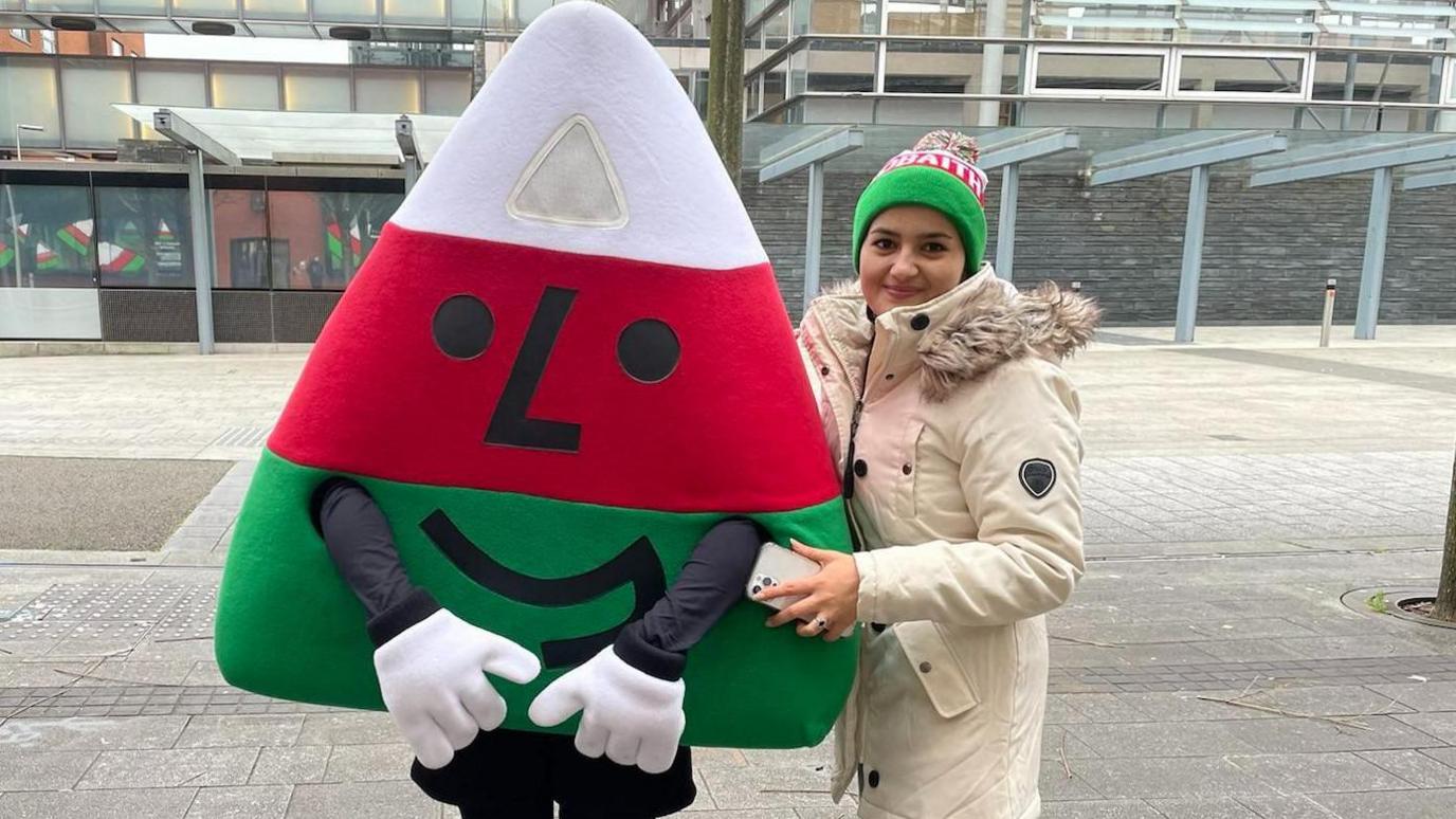 Mah in a large cream coloured coat and green & white beanie posing with the Urdd's Mascot - which is a triangle with red, white and green stripes and face on it in Cardiff Bay - outside the Senedd. 