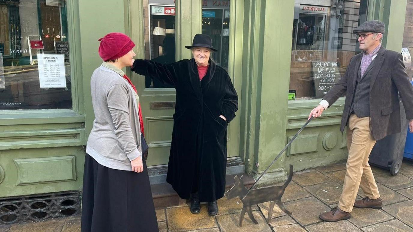 Three people standing in the doorway. The woman to the left is wearing a red hat, the woman in the centre has a black hat and the man on the end has a grey cap. 