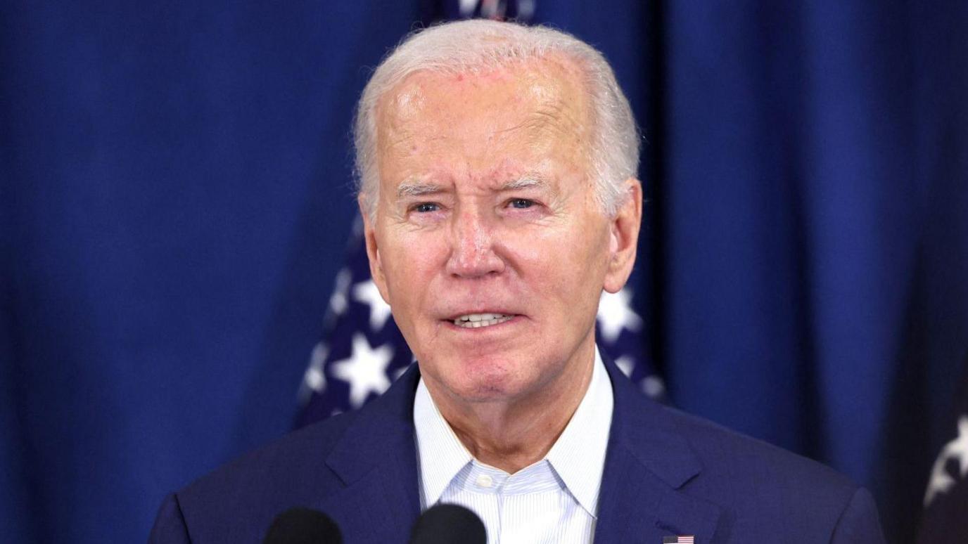 Joe Biden wears a navy suit as he addresses the media at a press conference. He is wearing a navy suit and white striped shirt with a US flag pin on his left lapel.