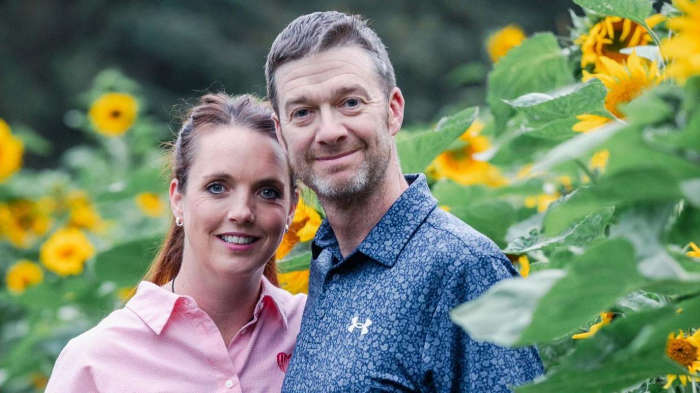 A man and woman pose for the camera and smile while standing in a field of sunflowers.