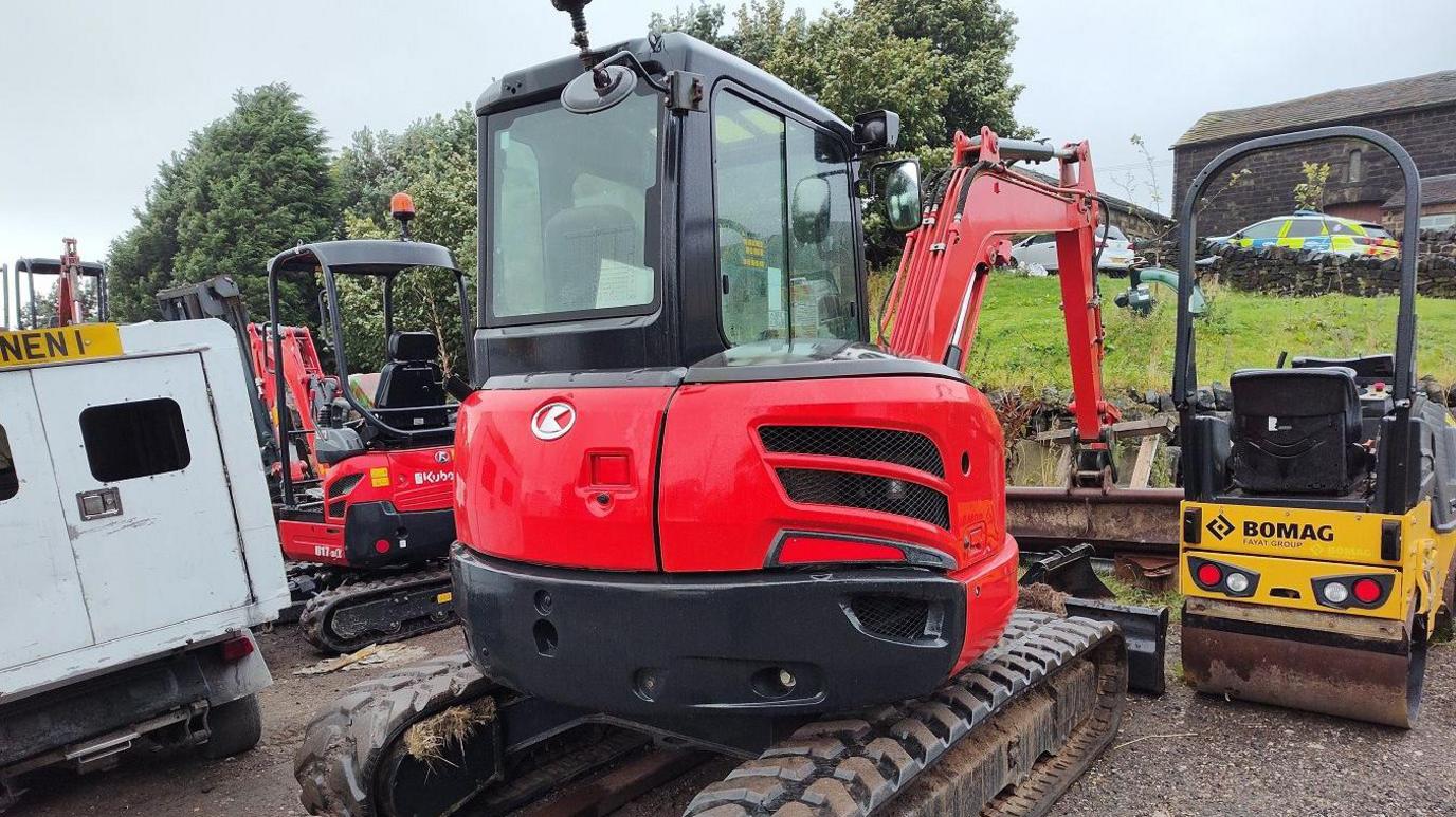 Two bright red diggers stand beside a small yellow compaction roller and a white van in a yard