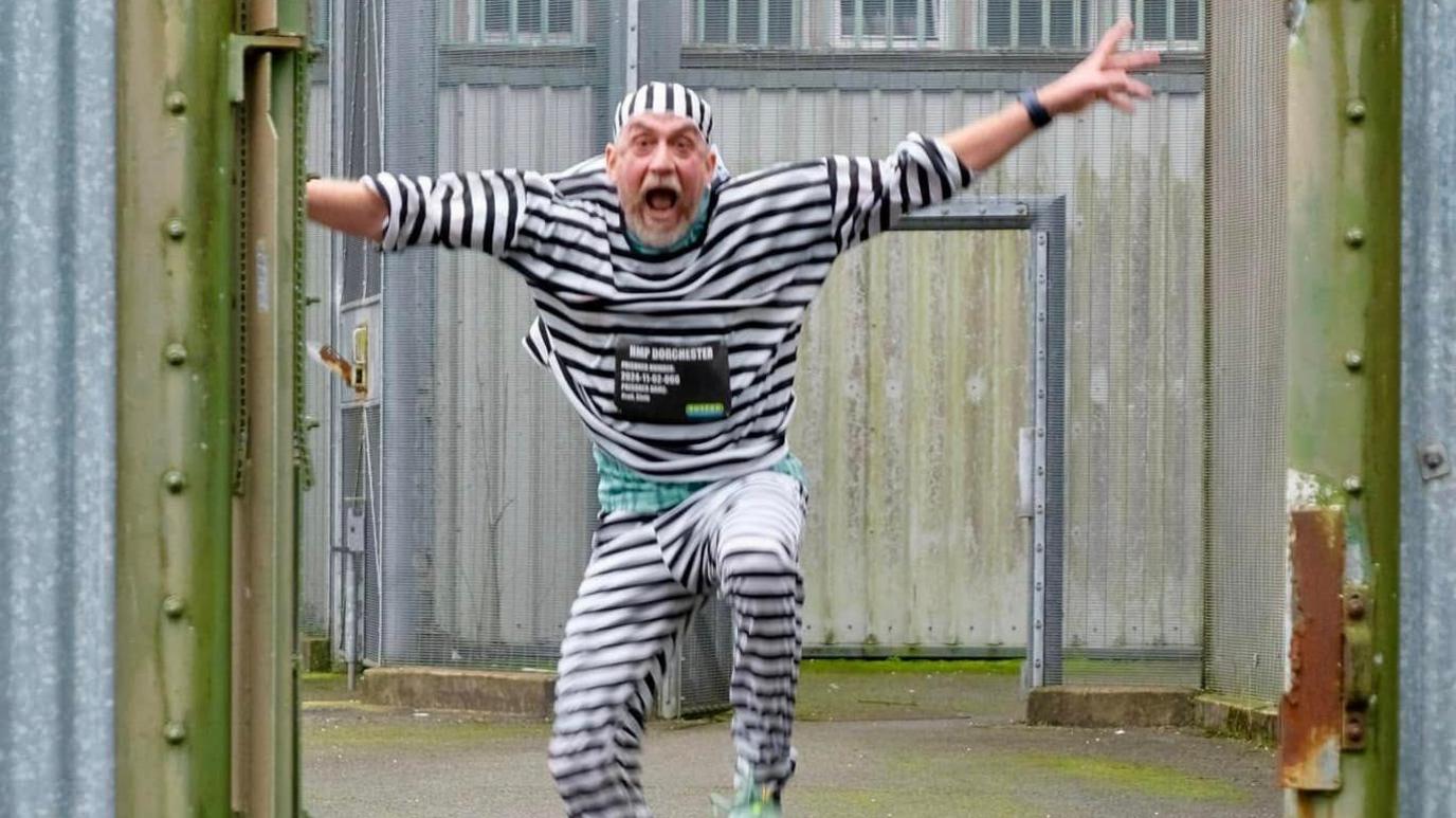 A runner dressed in black and white striped prison uniform leaping in the air as he runs towards the camera during the Dorchester Prison marathon. His arms are outstretched and his mouth and eyes are open wide.