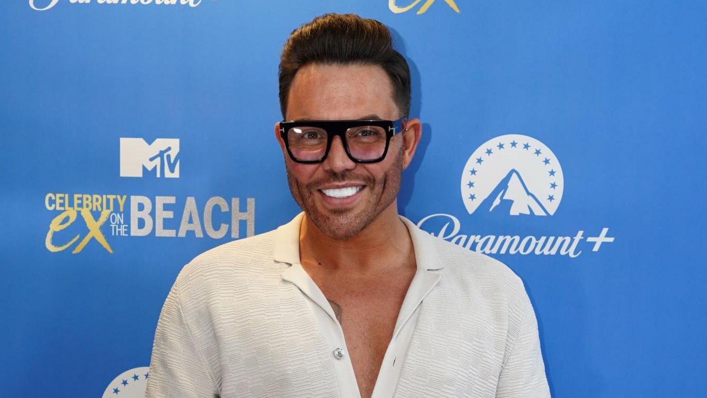 A smiling Bobby Norris looks directly at the camera, dressed in a white shirt and wearing large black-framed glasses. 