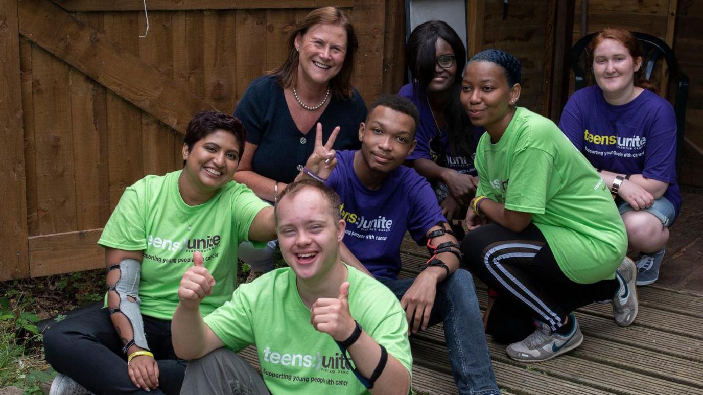 A woman and six young people sitting and crouching outside a wooden shed. The woman is wearing a navy top, three of the young people are wearing green T-shirts with the words "Teens Unite" on the front, and the other three are wearing purple T-shirts with the words "Teens Unite" on the front.