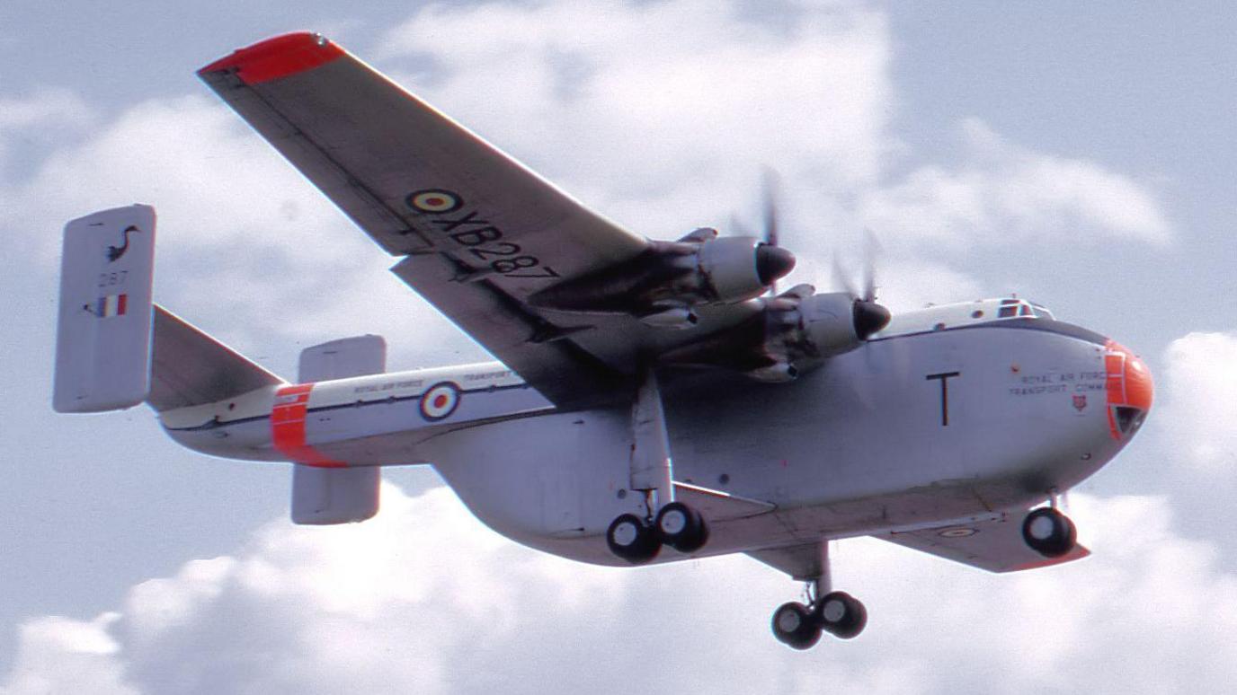 A large grey plane with a red nose and wing tips flies. It has four engines (two on each wing) and two vertical tail fins