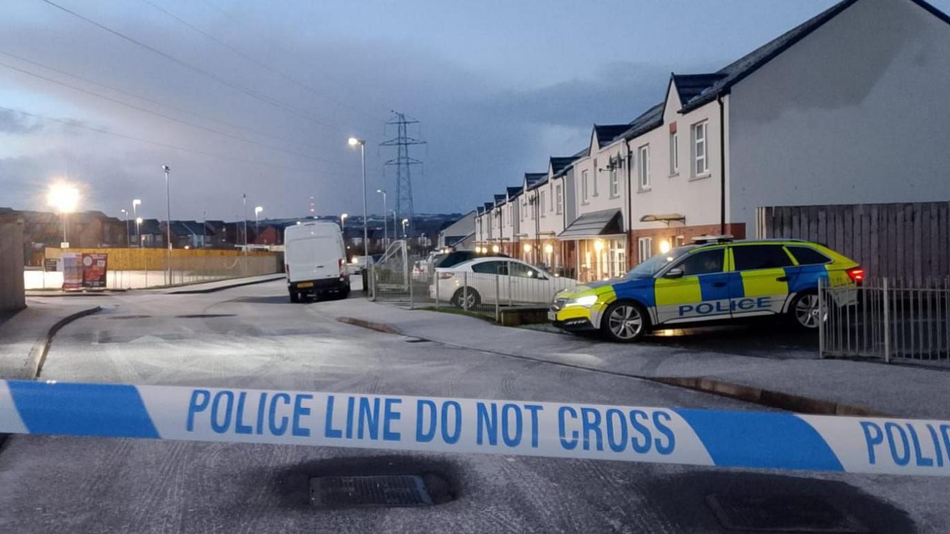 Police tape is in the foreground of the image, behind the tape sits a parked police car, which is in front of a row of houses. A white van and a number of parked car are on the road. the road is icy.