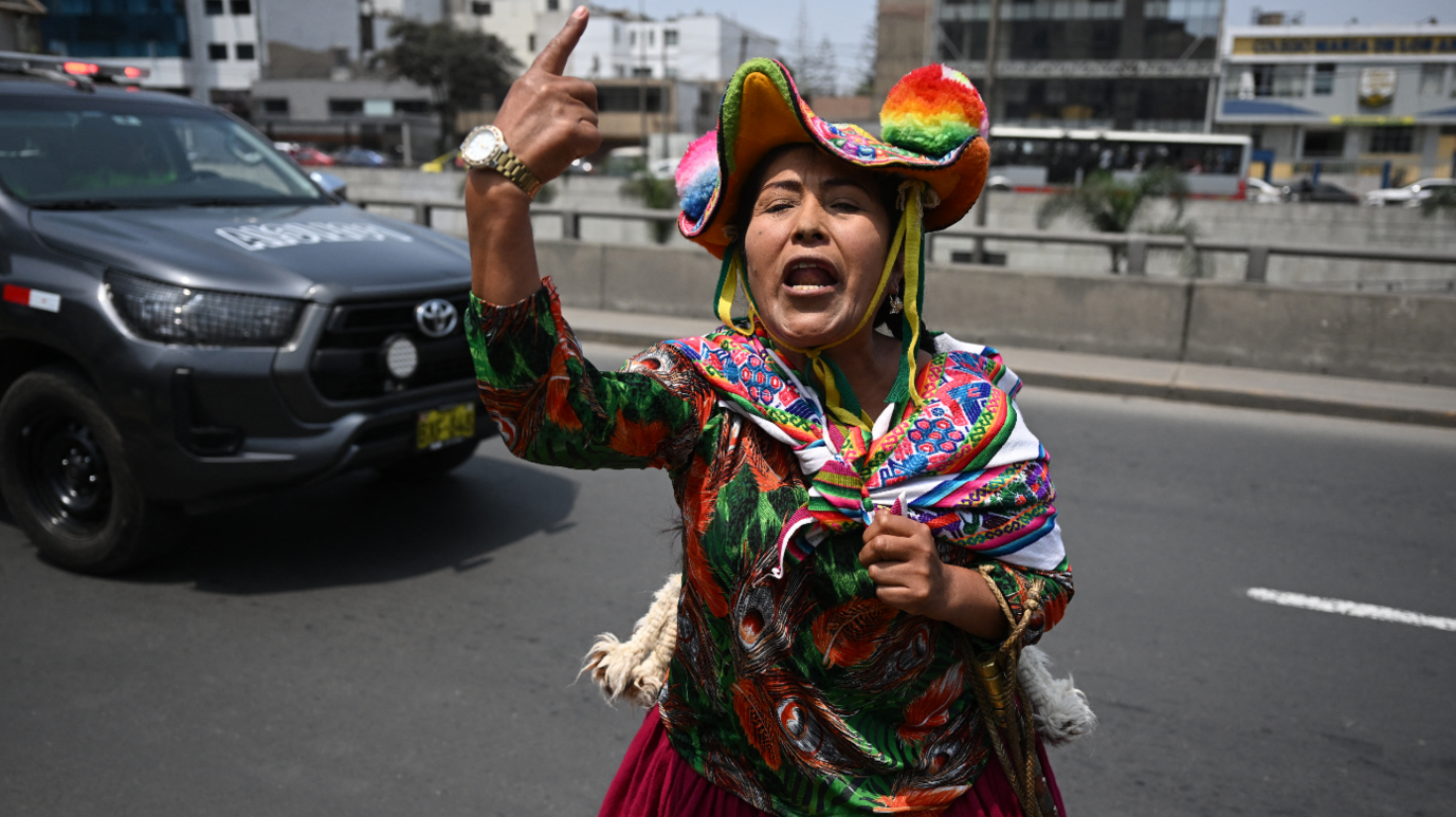 An indigenous woman wearing a colourful hat and clothing raises her index finger and shouts against the former president Fujimori. A Toyota vehicle is pictured in the background