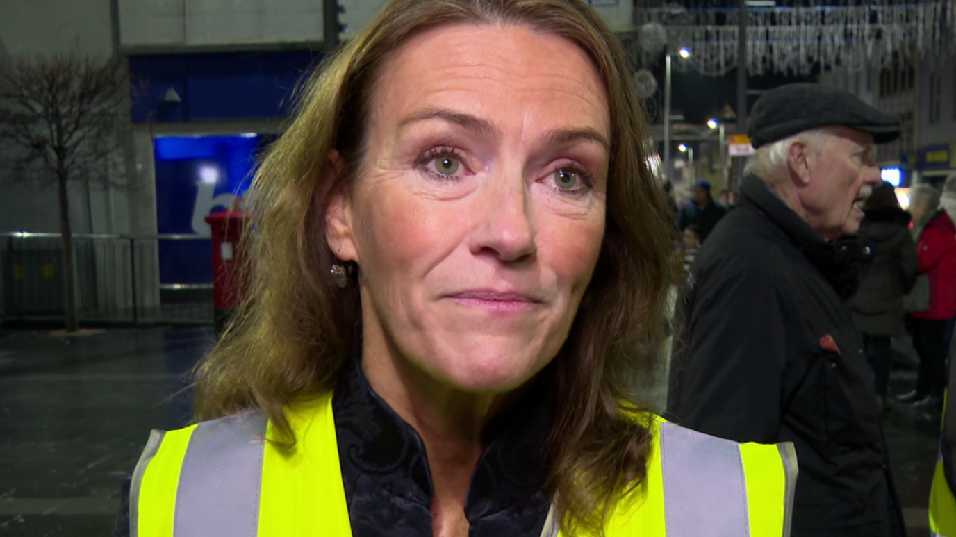 Helen O'Sullivan is wearing a yellow high viz jacket. She has wavy brown hair. Other people are standing behind her.