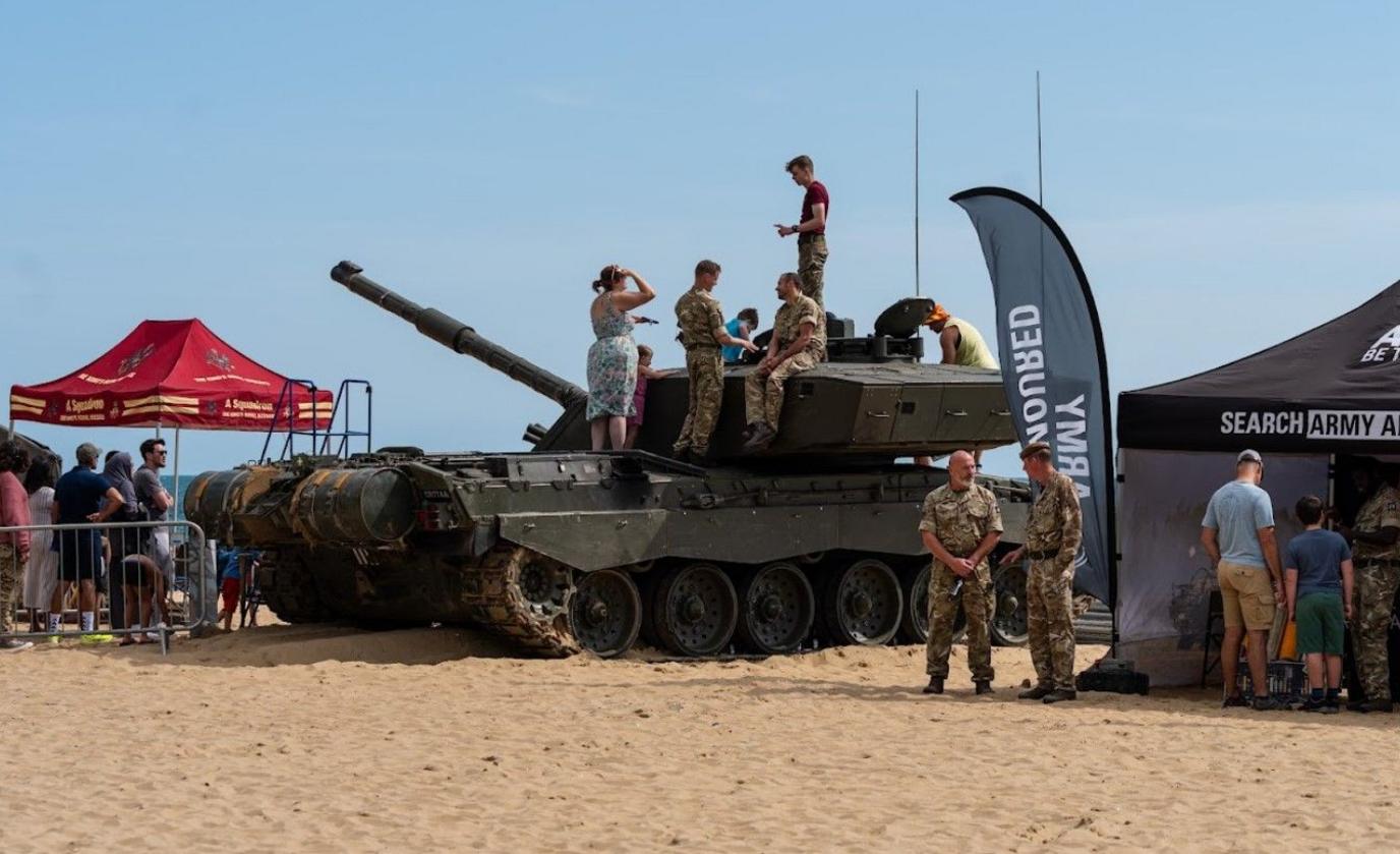 Army personnel in camouflage uniforms and members of the public standing on a tracked tank on Bournemouth beach